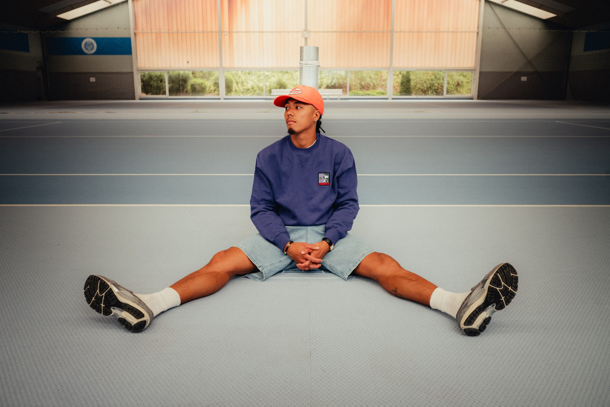 COLORBLND Model in blauem Sweater und Cap auf Indoor-Tennisplatz sitzend